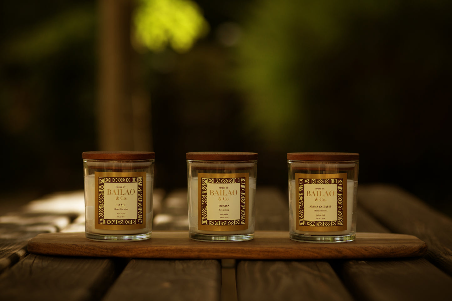 Three glass jar candles with wooden lids and decorative labels sit on a wooden board outdoors on a rustic wooden table, with a softly blurred green and brown background.