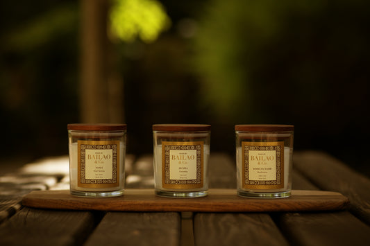 Three glass jar candles with wooden lids and decorative labels sit on a wooden board outdoors on a rustic wooden table, with a softly blurred green and brown background.