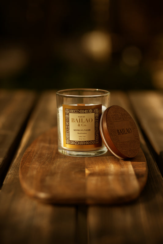 A glass jar candle with a gold label sits on a wooden board next to its round wooden lid, set on a rustic wooden table with a warm, dark bokeh background.