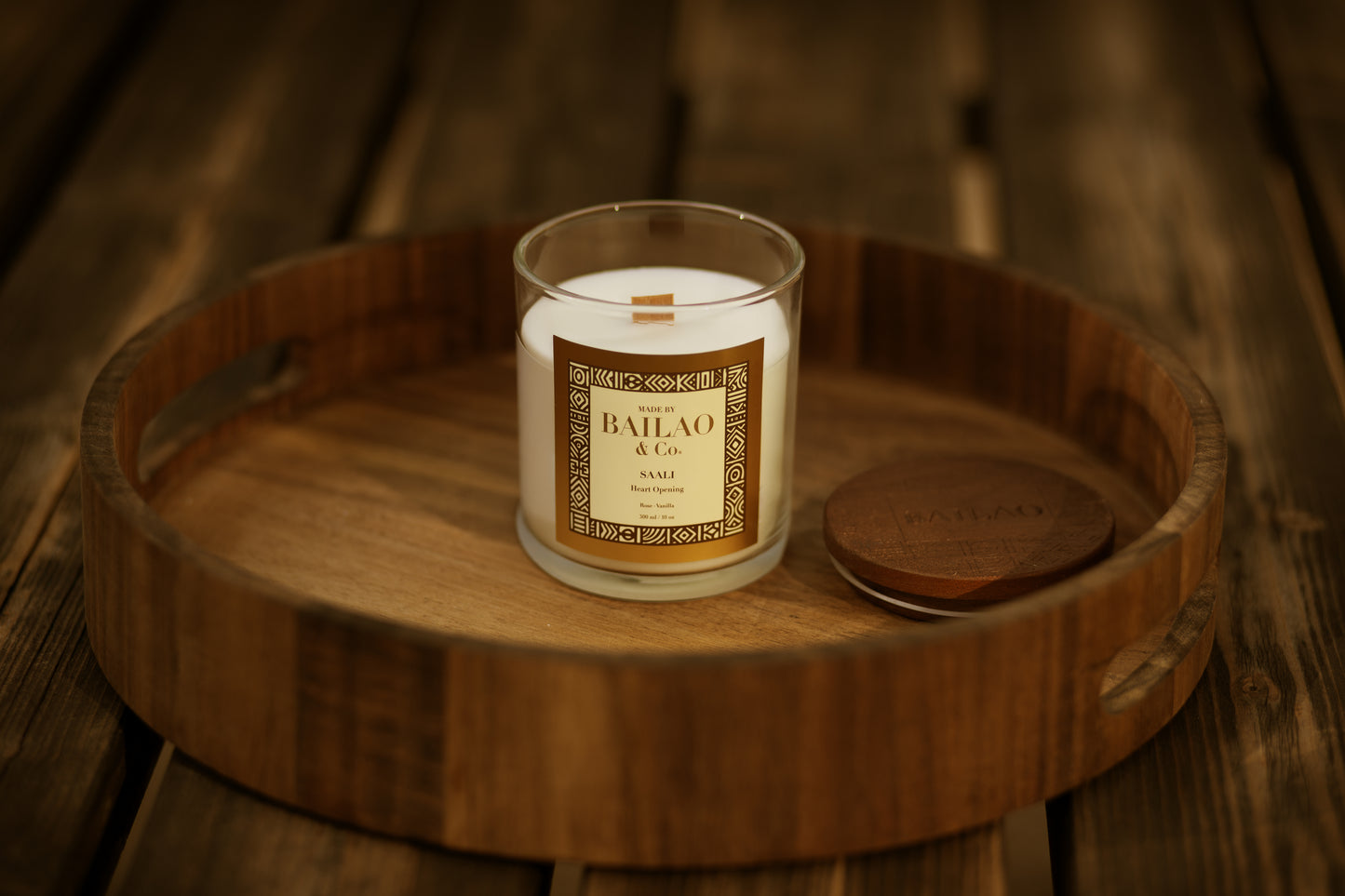 A glass jar candle with a cream-colored wax and a gold label reading 'BAHAO & CO' sits on a round wooden tray, with its matching wooden lid placed beside it on a rustic dark wood background.