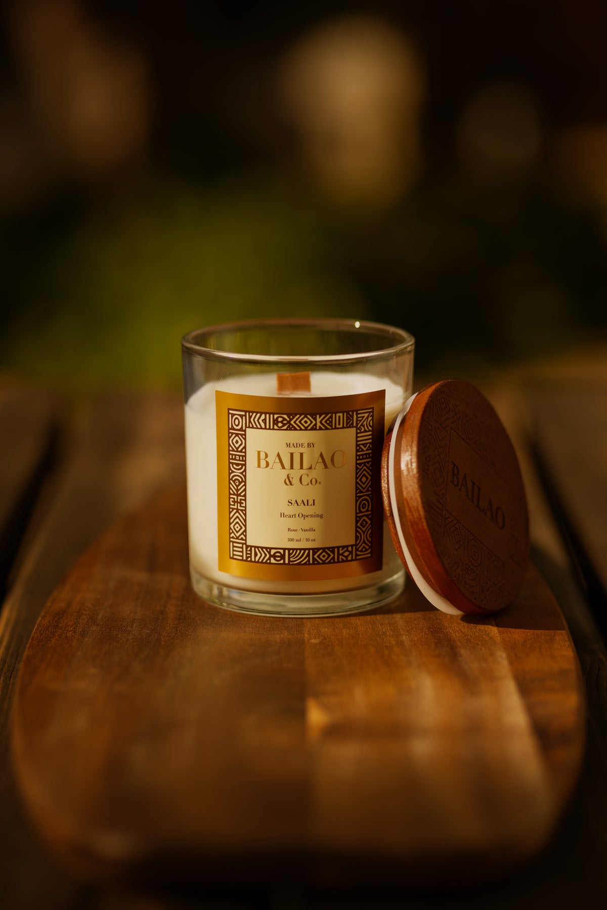 A Bailel & Co. scented candle in a clear glass jar with a cream-colored wax and a gold label, shown next to its round brown wooden lid on a rustic wooden board with a warm, blurred background.