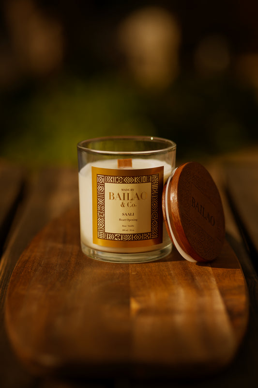A Bailel & Co. scented candle in a clear glass jar with a cream-colored wax and a gold label, shown next to its round brown wooden lid on a rustic wooden board with a warm, blurred background.